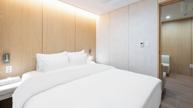 Habitación de hotel con una cama grande, paneles de madera en las paredes, una mesita de noche y un termostato en la pared, además de una puerta que da a otra habitación.