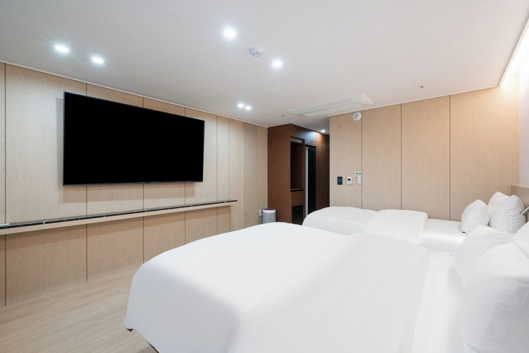 A hotel room with a large flat-screen TV mounted on the wall, a bed with white bedding, and a wooden panel wall.