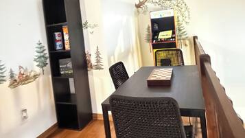 A dining area with a wooden table, black chairs, a black bookshelf, and a wall-mounted arcade game.