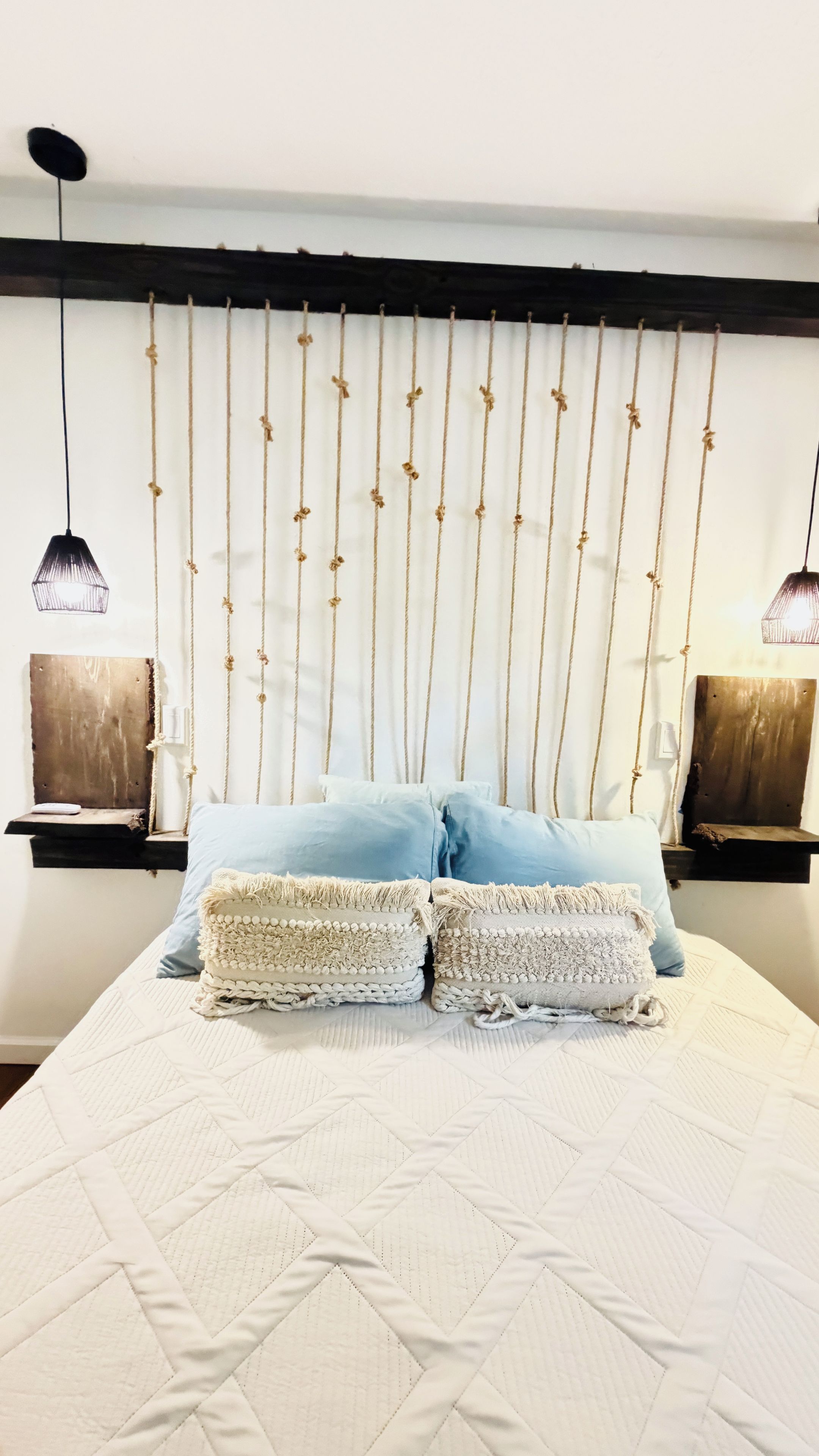 A bed with a white quilted bedspread and two light blue pillows, a wooden headboard with hanging ropes, and two bedside tables with lamps.