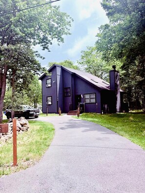 A two-story house with a dark exterior, a chimney, and a paved driveway leading to the entrance. - Graphite Grove - Arcade, Fire Pit & Covered Deck - Pet Friendly - Poconos (Tobyhanna)