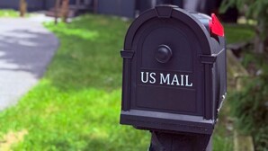 Ein Briefkasten mit der Aufschrift 'US MAIL', ein Haus im Hintergrund und ein klarer Himmel.