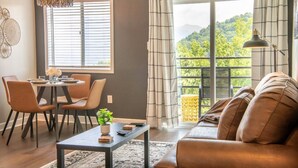 Ein Wohnzimmer mit einer Couch, einem Esstisch, Stühlen und Blick auf Grün hinter großen Fenstern.
