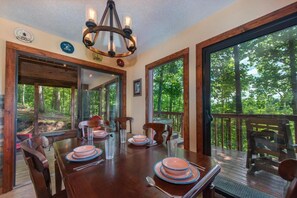 A dining area with a wooden table set for four, large windows, and a view of the forest. - Three Little Bears (Gatlinburg)