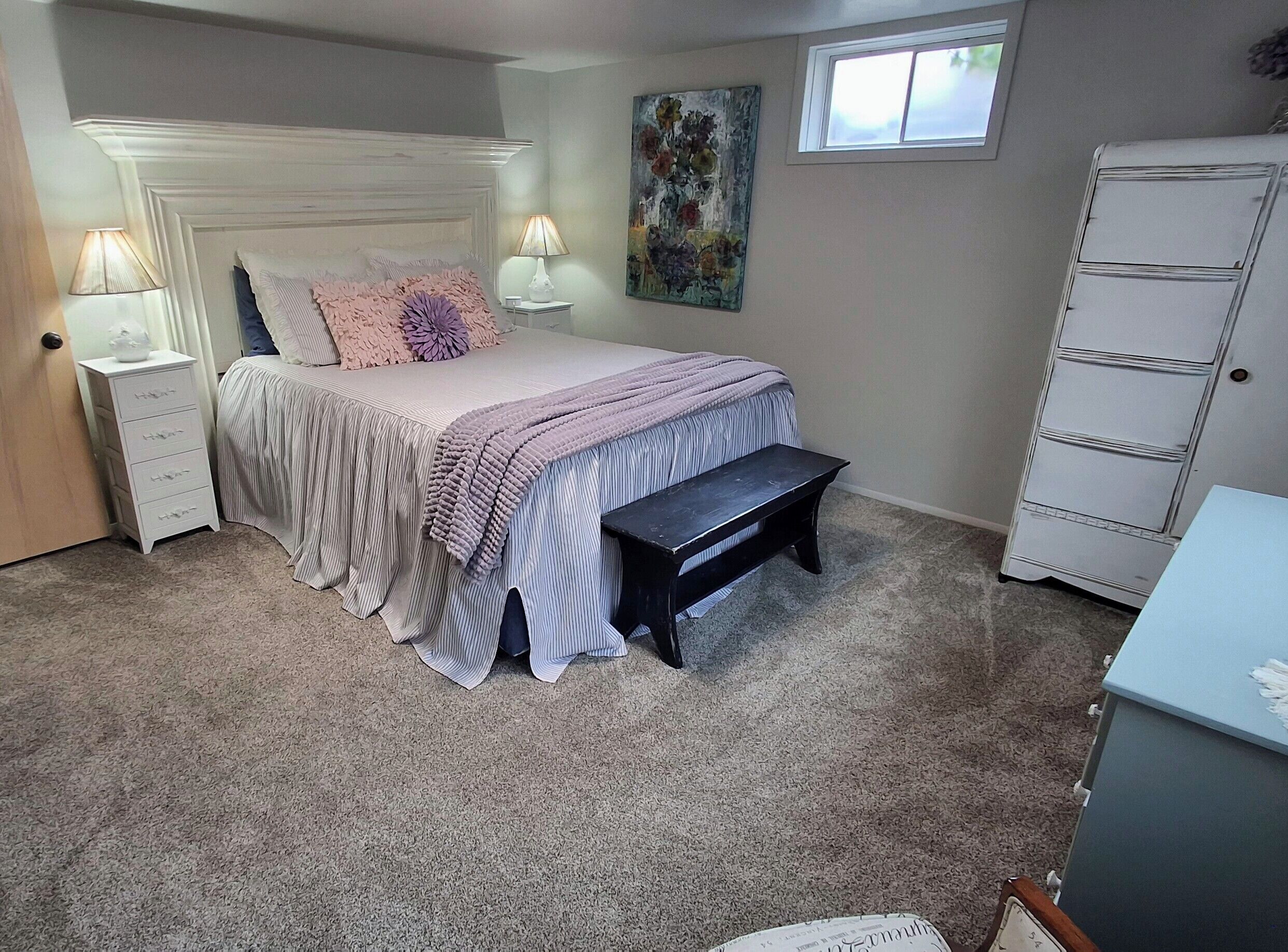 A bedroom with a bed, bedside lamps, a bench, a dresser, and a painting on the wall.