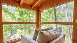 Interior de una cabaña de madera con amplias ventanas, un sofá y vistas a un bosque.