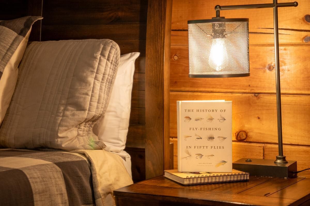A bed with pillows, a book titled 'The History of Fly-Fishing in Fifty Flies,' and a lamp on a wooden nightstand.
