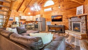 Interior de una cabaña de troncos con sala de estar, cocina, chimenea y techo de madera.