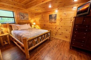 Quarto de cabana de madeira com cama, mesa de cabeceira, abajur, cômoda e obras de arte nas paredes.