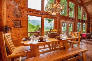 A wooden cabin interior with a dining table set for four, large windows, and a deer head mounted on the wall. - Simply Incredible (Sevierville)