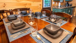 Een houten eettafel gedekt met borden, glazen en bestek in een kamer in chaletstijl, met houten wanden en een keuken op de achtergrond.