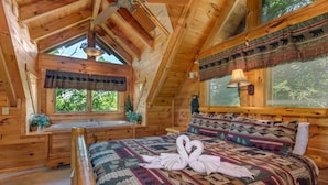 Un dormitorio de cabaña rústica con una cama grande, una ventana con vistas a los árboles, techo de madera y una ventana con cortina estampada.