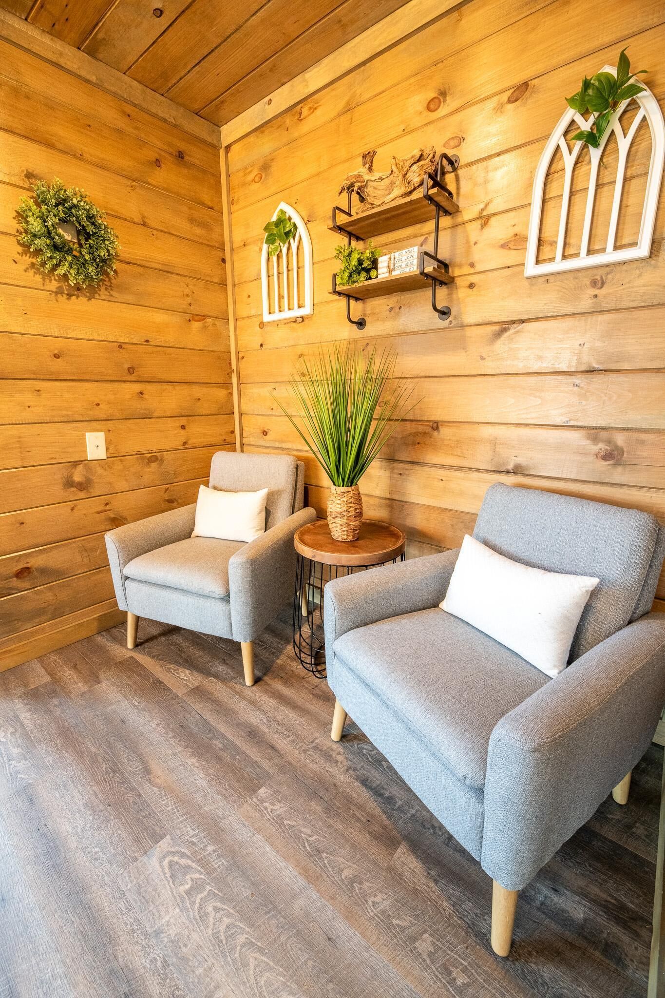 A cozy room with wooden walls, two grey armchairs, a small round table with a plant, and decorative wall shelves.