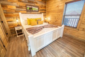 A wooden-paneled bedroom with a bed, bedside tables, a nightstand, a window with blinds, and a mounted picture. - Amazing Grace (Sevierville)