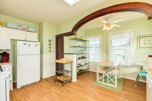Eine Küche mit einem weißen Kühlschrank, einer Kaffeemaschine und einer Mikrowelle. Es gibt einen Holztisch mit einem Stuhl, ein Regal mit Gläsern und einen Deckenventilator.