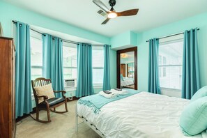 Ein Zimmer mit einem Bett, einem Schaukelstuhl, einem Deckenventilator und großen Fenstern mit Vorhängen.