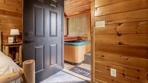 Un interior de madera con una puerta que da a una habitación con cama, una mesita de noche con lámpara y un banco de madera.