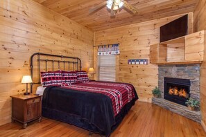 A log cabin bedroom with a bed, nightstand, lamp, window with blinds, and a stone fireplace. - Splash Mountain (Sevierville)