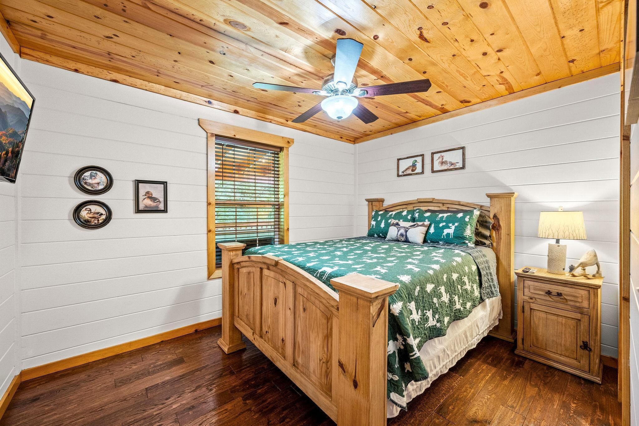 Un dormitorio con ventilador de techo de madera, cama con colcha estampada verde, mesita de noche con lámpara y ventana con persianas.