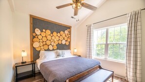 Een slaapkamer met een bed, nachtkastjes, een bankje, een plafondventilator, een raam met gordijnen en een kunstwerk aan de muur.