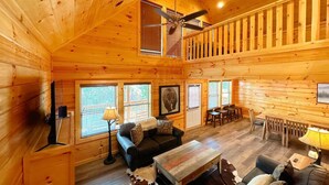A log cabin interior with a ceiling fan, wooden walls, a black sofa, a wooden coffee table, and a dining area with a table and chairs. - Shepherd's Rest (Pigeon Forge)