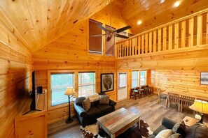A log cabin interior with a ceiling fan, wooden walls, a black sofa, a wooden coffee table, and a dining area with a table and chairs. - Shepherd's Rest (Pigeon Forge)