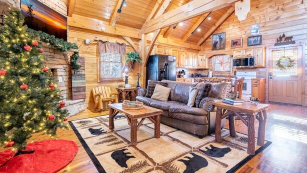 A cozy log cabin interior with a Christmas tree, wooden furniture, and a stone fireplace. - Smokey's Hideaway (Sevierville)