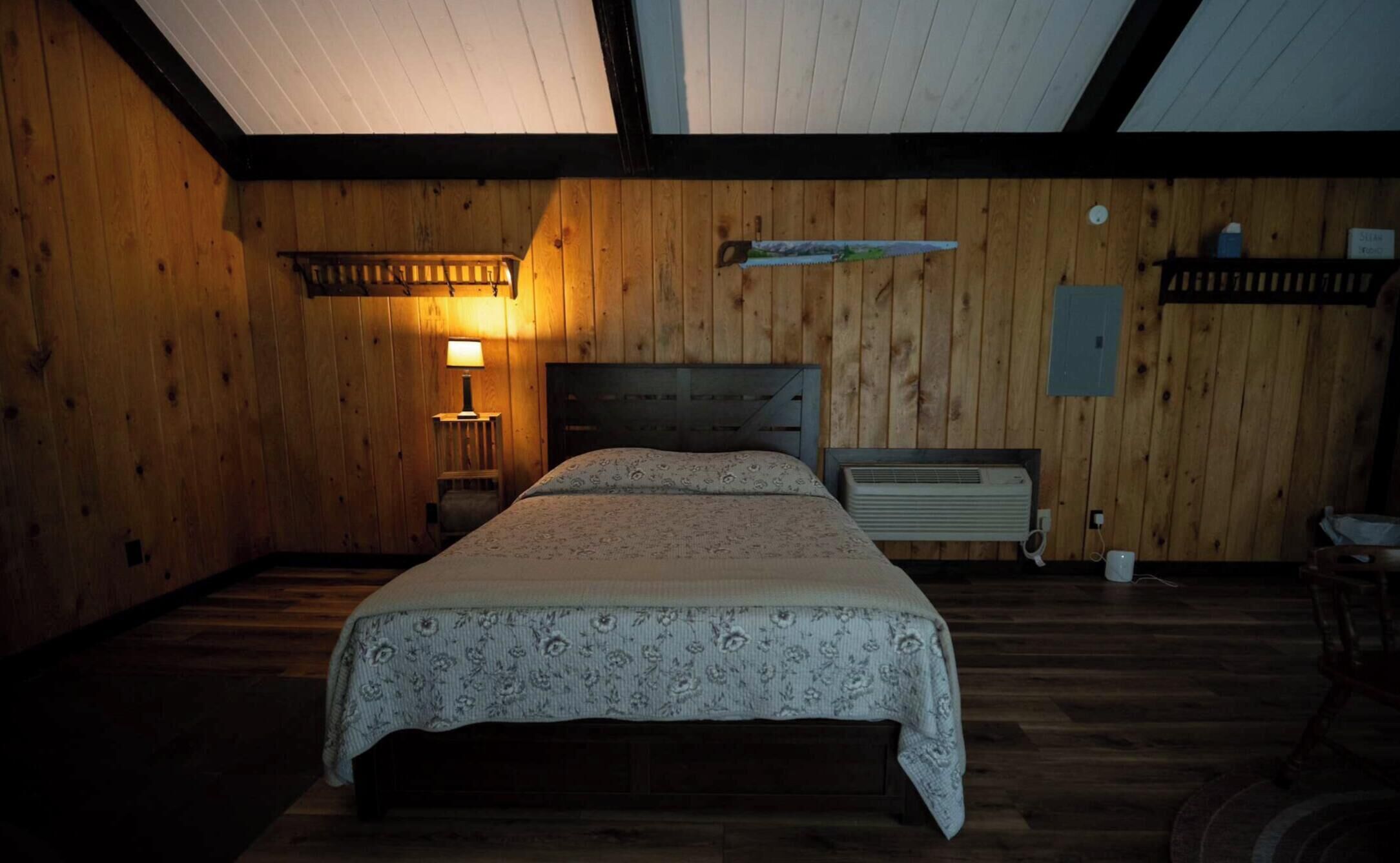 Une pièce aux murs en bois, un lit recouvert d’une couvre-lit à motifs, une table de chevet avec une lampe et un radiateur fixé au mur.
