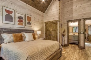 Ein Schlafzimmer mit einem großen Bett, Holzwänden und einer Holzbalkendecke. Ein Badezimmer ist durch eine offene Tür sichtbar.