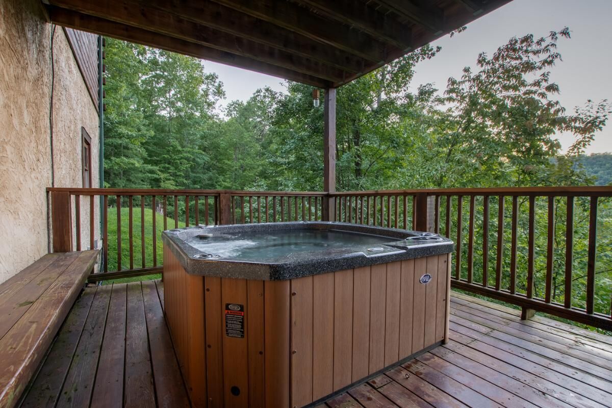 A wooden deck with a hot tub, wooden benches, and a railing overlooking a forested area.