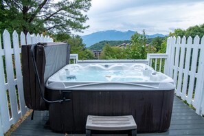 A hot tub on a wooden deck with a white picket fence, surrounded by greenery and mountains in the background. - Ski View Summit (Gatlinburg)