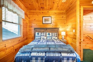 Ein Zimmer in einer Holzhütte mit einem Bett, einem gerahmten Bild an der Wand und einem Fenster mit Jalousien.
