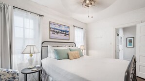 Un dormitorio con una cama, dos mesitas de noche, una mesita auxiliar, un ventanal con cortinas, un cuadro en la pared y una puerta.