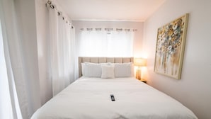 A hotel room with a large bed, a bedside table with a lamp, a painting on the wall, and sheer curtains on the windows. - The Jeffrey Martin (Gatlinburg)