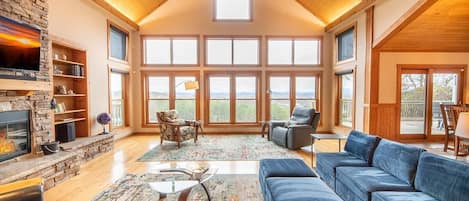 A spacious living room with a stone fireplace, large windows, a blue sofa, and a high ceiling with wooden beams.