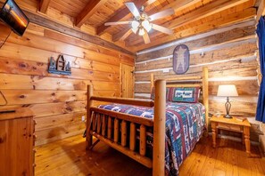 A log cabin bedroom with a wooden bed, a ceiling fan, a small table with a lamp, and a wall-mounted cross. - Mountain Memories 2 (Sevierville)