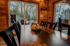 A wooden interior with a dining table, chairs, a bowl of fruit, and a view of the outdoors through large windows. - Z - Highland Haven (Sevierville)