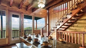 A wooden interior with a dining table set for four, a staircase, and large windows offering a view. - Windows to Heaven (Sevierville)