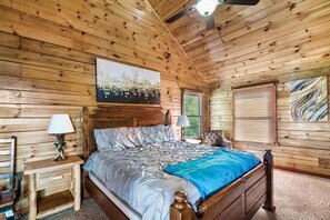 A wooden-paneled room with a bed, two nightstands, a ceiling fan, and a window with blinds. - Windows to Heaven (Sevierville)