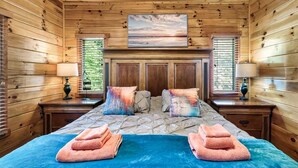 A wooden-paneled room with a bed, two nightstands, and a painting above the bed. - Windows to Heaven (Sevierville)