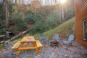A wooden picnic table with chairs, a fire pit, and a grill in a forested area. - Three Little Bears 2 (Gatlinburg)