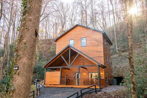 A log cabin with a porch, a small attached structure, and a forested backdrop. - Three Little Bears 2 (Gatlinburg)