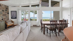 A high-ceilinged room with a stone wall, a sofa, a dining table with chairs, and large windows offering a view of the beach. - The Beach Bungalow | Coastal Vibe | Private Beach! (West Olive)