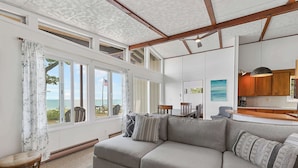 A modern living room with a large gray sofa, a dining table, and a kitchen area. - The Beach Bungalow | Coastal Vibe | Private Beach! (West Olive)