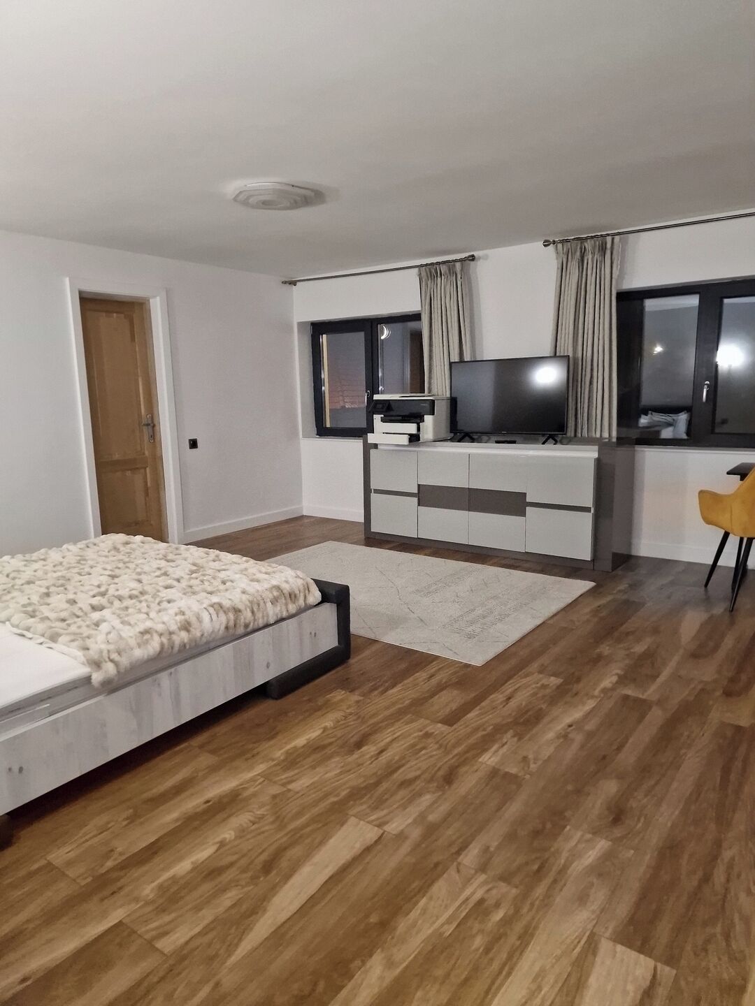 Ein Schlafzimmer mit einem Bett, einem Schreibtisch mit Fernseher, einem Sessel und einem Fenster mit Vorhängen.