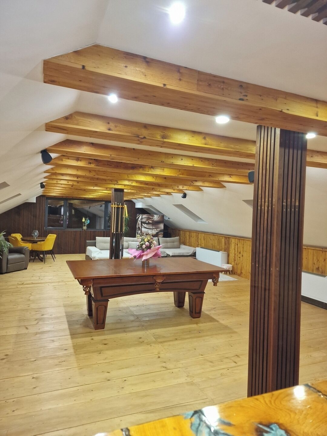 A loft-style room with wooden beams, a dining table, sofas, and a large window.