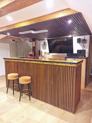 A bar area with a wooden bar, two stools, and a TV screen.