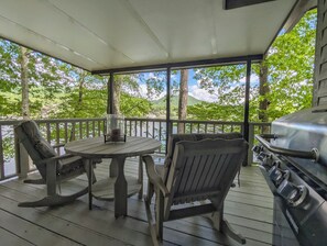 A covered outdoor area with a table and chairs, a grill, and a view of a lake and trees. - Llyn Blaen House at Lake Junaluska (Lake Junaluska)