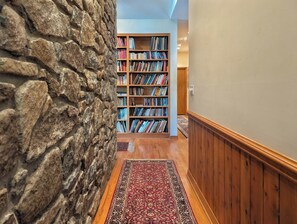 A hallway with a stone wall, wooden flooring, a patterned rug, and a bookshelf filled with books. - Llyn Blaen House at Lake Junaluska (Lake Junaluska)