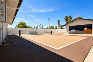 An outdoor tennis court with a net, surrounded by a white fence, palm trees, and a building with a mural. - Ultimate Retreat|Pool~Spa~Pickleball~GameRoom~BBQ (Twentynine Palms)
